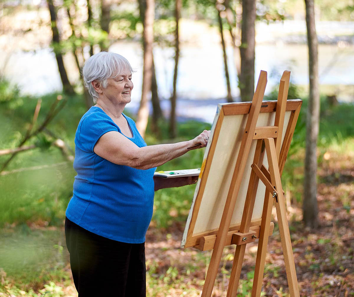 A senior woman stands outside in a nature setting painting on a easel