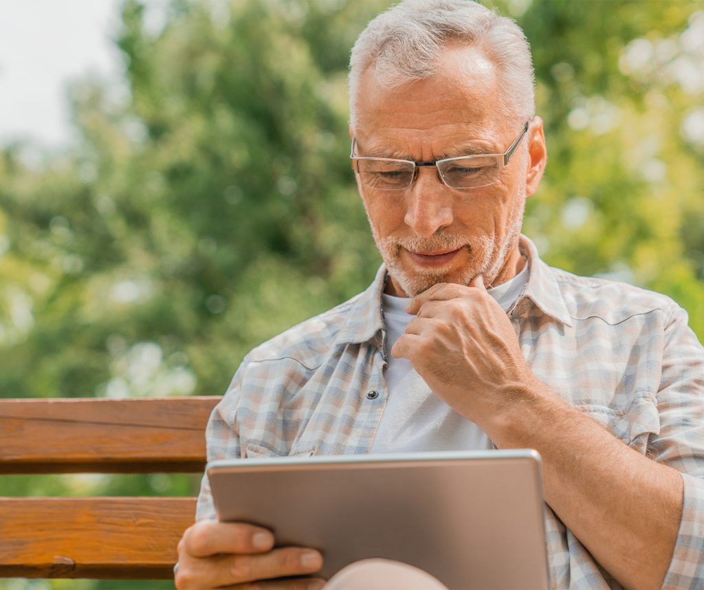older man sitting on a bench outside and reading an ipad