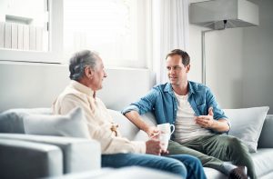younger man talking to elderly parent about changes while sitting on couch