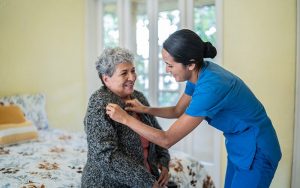 Mid adult nurse helping senior woman dress shirt in bedroom at nursing home