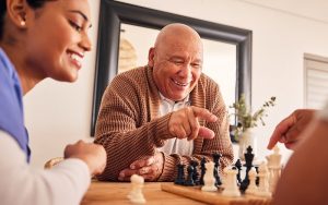 Senior man, nursing home and chess for game, competition and happy with friends, strategy and relax together. Elderly person, nurse and board for contest with mindset, excited smile or point at table