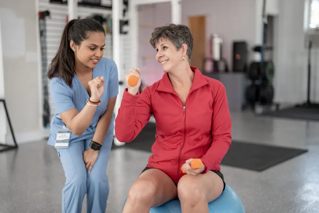 A resident woman is discovering what is occupational therapy for seniors at Rolling Green Village in Greenville SC. The elderly woman is lifting weights while balancing on ball with encouragement from a occupational therapist.