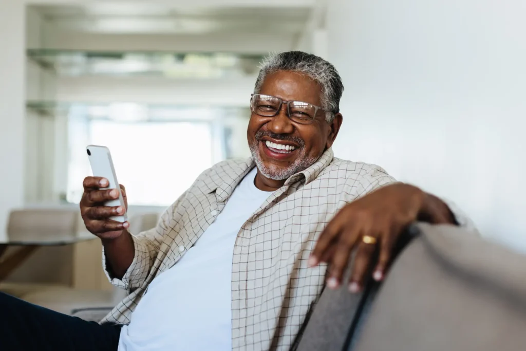 An older man on couch smiling at camera holding a cell phone to look at Buy-in vs Monthly rental communities for retirement in Greenville, SC.