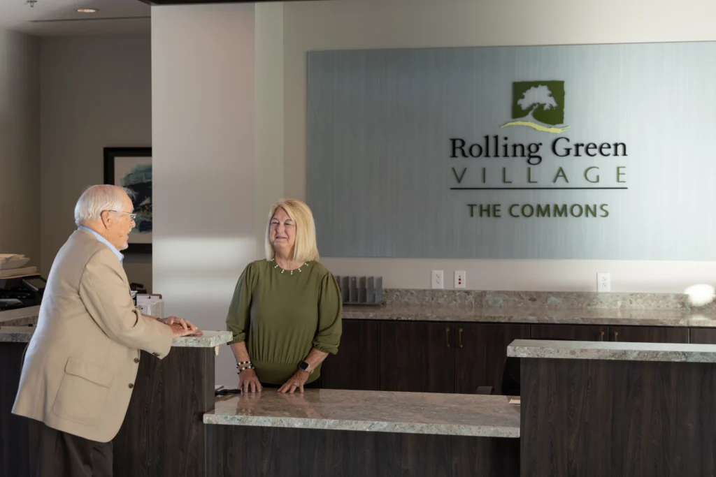 A senior man talking with a receptionist at Rolling Green Village asking about the life plan community expansion benefits in Greenville, SC in 2026.