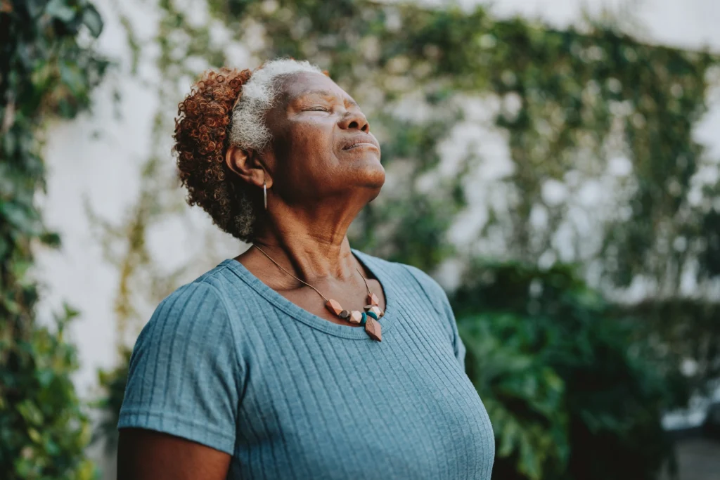 A senior woman with eyes closed taking a deep, mindful breath in a lush garden, representing spiritual and environmental aspects of the 8 dimensions of wellness for seniors.