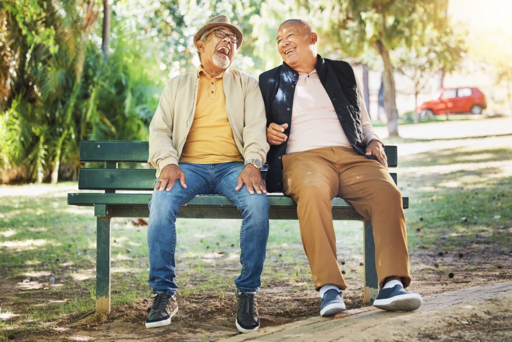 Two senior men laughing joyfully while sitting on a park bench, highlighting social and emotional health as part of the 8 dimensions of wellness for seniors.