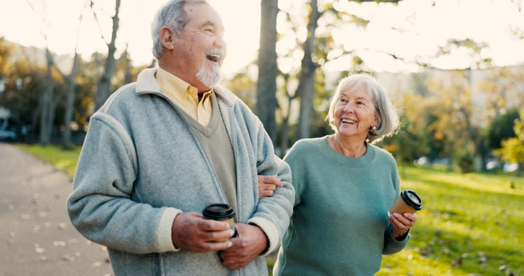 A happy senior couple walking through a sunlit park with coffee, illustrating physical and social activity within the 8 dimensions of wellness for seniors.