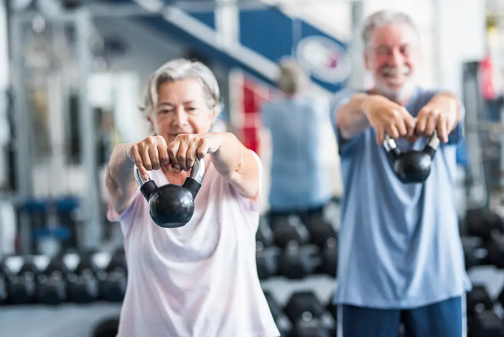 Seniors at Rolling Green Village practice kettlebell swings during a group exercise class in a gym setting, staying strong through weight lifting for seniors.