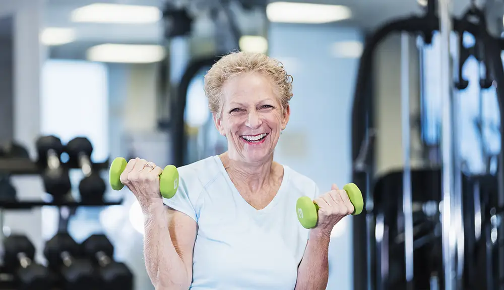 A senior resident at Rolling Green Village smiles while doing bicep curls with green dumbbells in a fitness room during a weight lifting for seniors workout.
